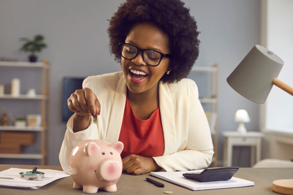Happy black woman puts coin in pink piggy bank. Smiling cheerful businesswoman sitting at office desk with calculator and money box. People and finance, successful business, saving up concept