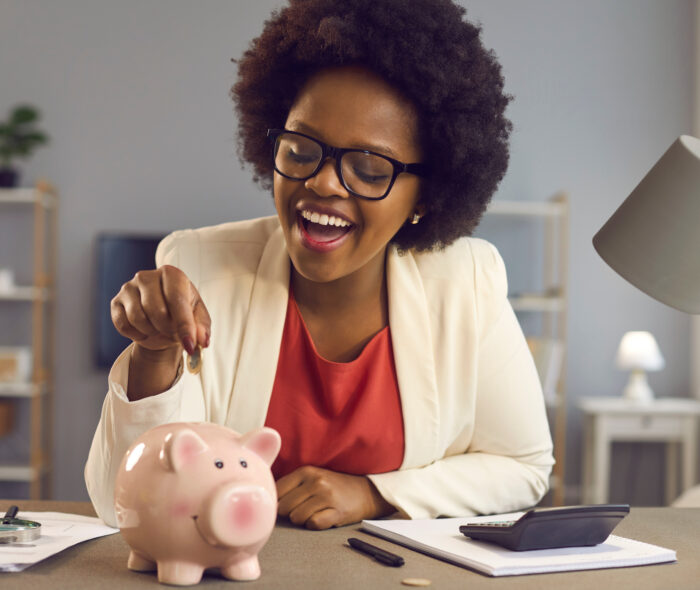 Happy black woman puts coin in pink piggy bank. Smiling cheerful businesswoman sitting at office desk with calculator and money box. People and finance, successful business, saving up concept