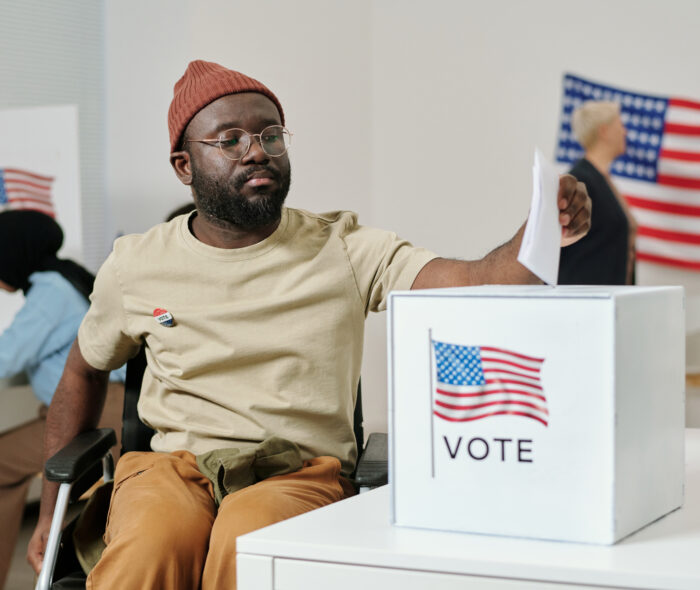 African American young man using a wheelchair and putting paper ballot paper into box with USA flag